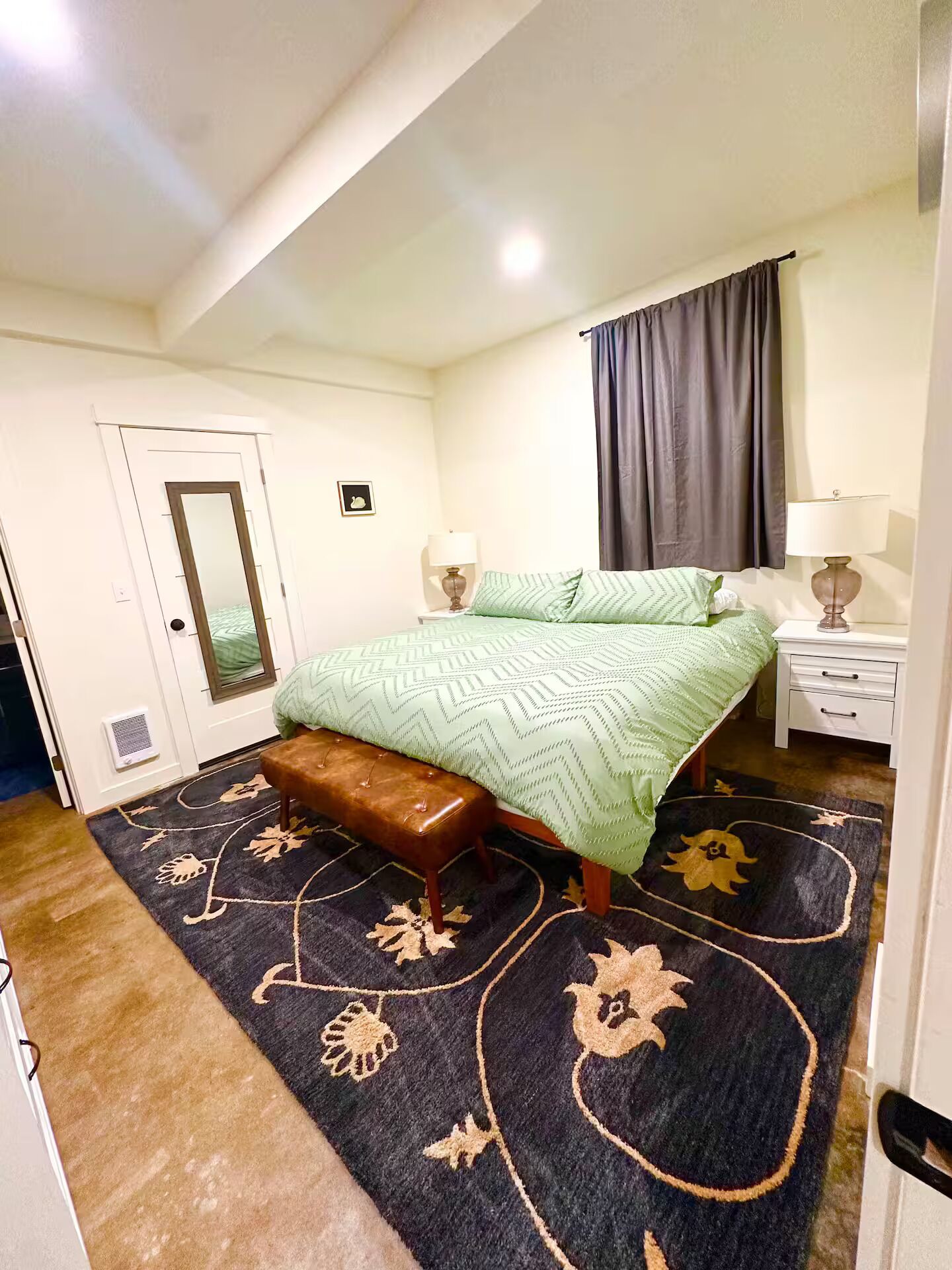 A bedroom with a bed, bench, bedside tables, a mirror, and a window with curtains.