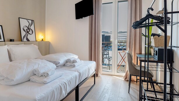 Quarto de hotel com cama, televisão, varanda com mesa e cadeiras, e um cabideiro.