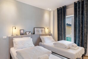 Habitación de hotel con dos camas, cabecera con lámpara, un cuadro enmarcado y una ventana con cortinas.