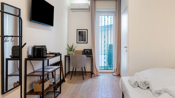 A compact hotel room with a bed, a desk with a chair, a flat-screen TV, a small table with a plant, and a window with curtains. - Giulietta Suite with Private Bathroom, Balcony, Wi-Fi, and Air Conditioning (Verona)