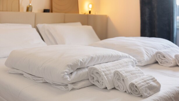 Un lit bien fait, avec du linge de lit blanc et des serviettes pliées, une tête de lit et une table de chevet avec une lampe.