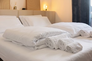 Un lit bien fait, avec du linge de lit blanc et des serviettes pliées, une tête de lit et une table de chevet avec une lampe.