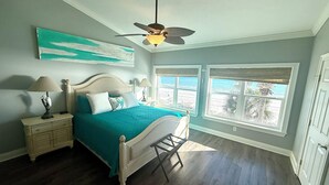 A bedroom with a bed, nightstands, a ceiling fan, and a view of the beach through large windows. - Gulfs Edge Townhome #16 - Sealee (Panama City Beach)