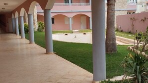 Une allée couverte bordée de colonnes, une pelouse verdoyante et un bâtiment aux murs roses et doté d’un balcon.