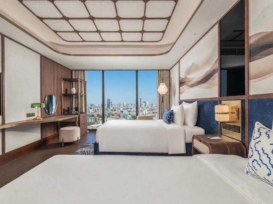 A modern hotel room with a large bed, a desk, a chair, a TV, and a city view.