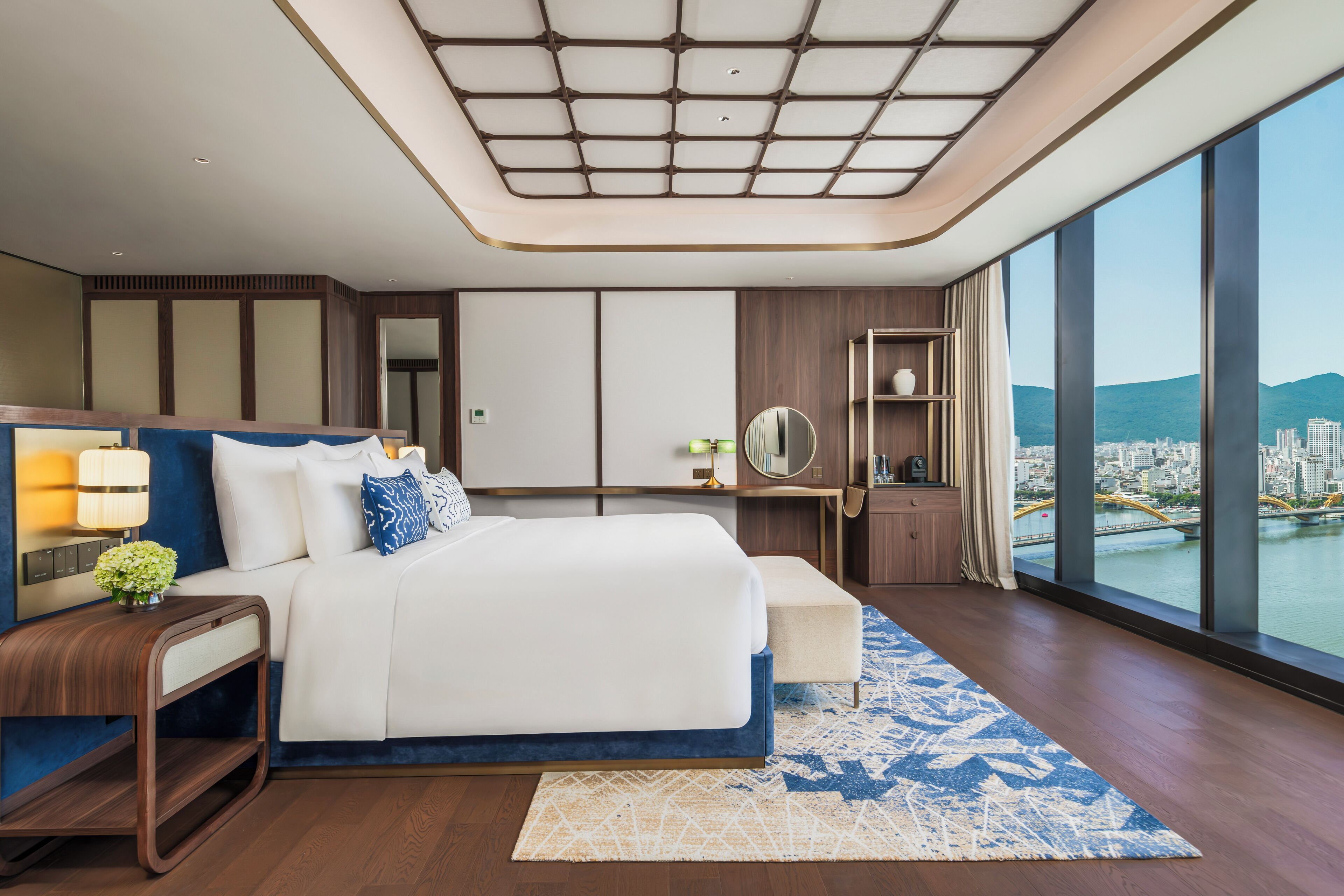 A modern hotel room with a large bed, a desk, a mirror, and a view of a cityscape.