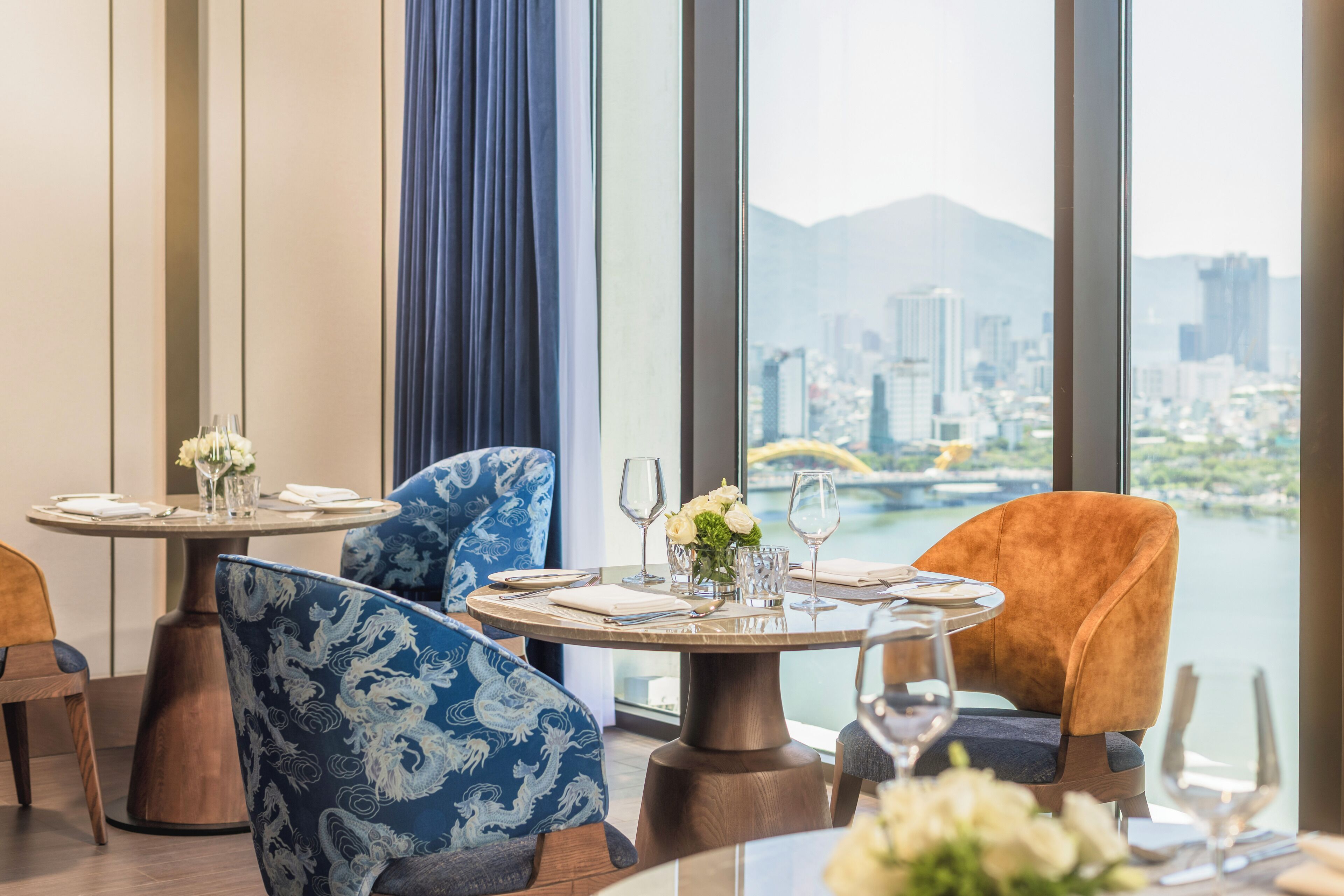 Un comedor con mesas redondas, sillas y vistas a un paisaje urbano a través de amplios ventanales.