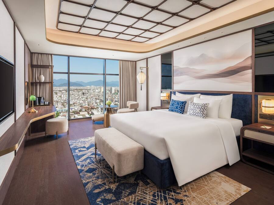 Quarto de hotel moderno com uma cama grande, uma escrivaninha, uma cadeira e vista para a paisagem urbana.