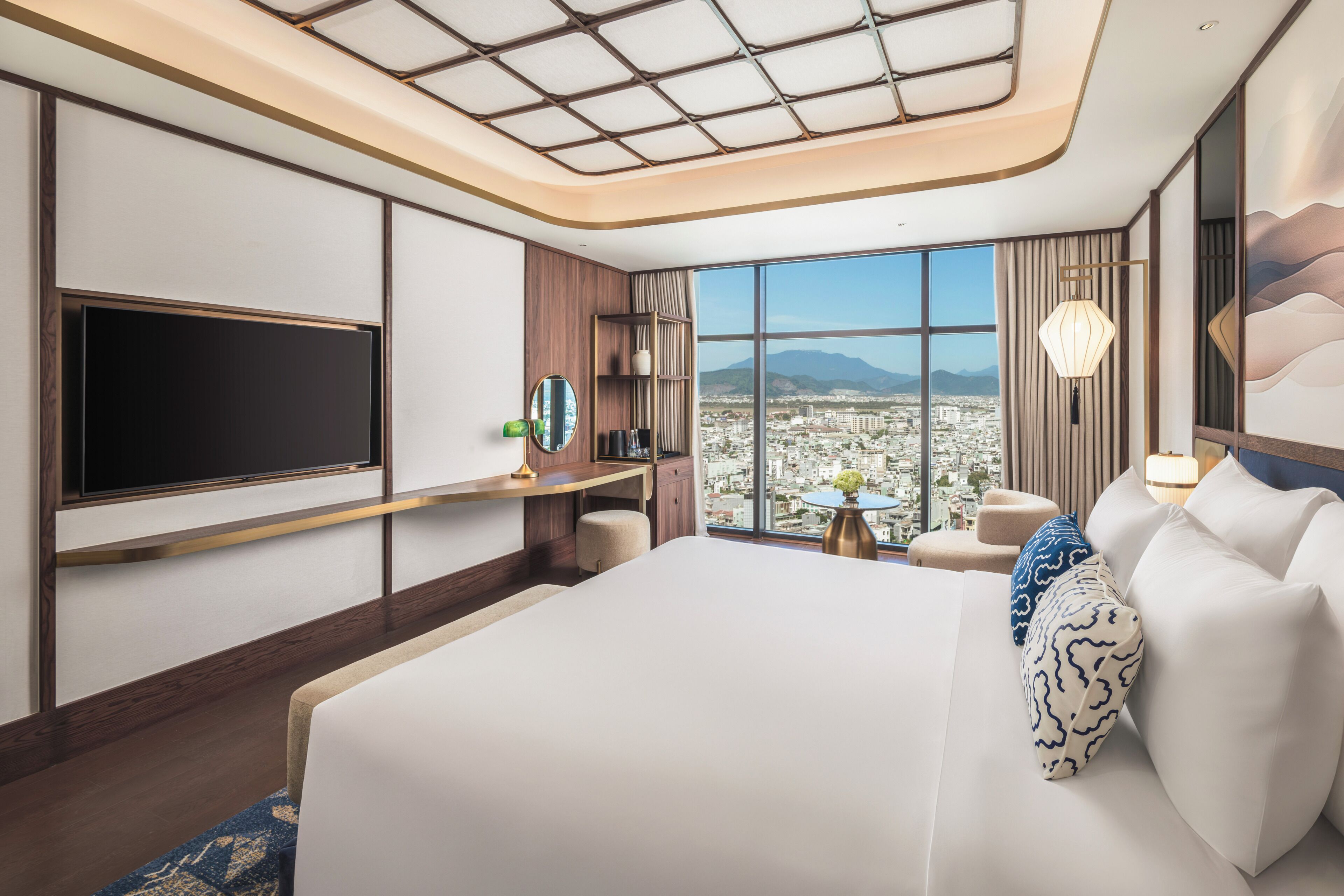 A hotel room with a large bed, a flat-screen TV, a desk with a chair, a small table with a vase, and a view of a cityscape through large windows.