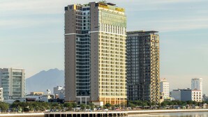 A high-rise building with multiple floors, a prominent sign at the top, and a reflection in the water in front. - TUI BLUE Han River (Da Nang)