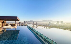 Une piscine à débordement située sur le toit, offrant une vue sur le paysage urbain, un pont et une chaîne de montagnes à l’horizon.