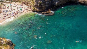 An aerial view of a beach with clear turquoise water, rocky cliffs, and a sandy beach area with people swimming and sunbathing. - Villa Eterea - Five Bedroom Villa, Sleeps 10 (Selva di Fasano)