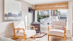Ein Wohnzimmer mit zwei Sesseln, einem Kamin, einem großen Fenster mit Blick auf Gebäude und einem Bild an der Wand.