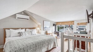 Ein Schlafzimmer mit schrägen Wänden, einem Bett mit gemustertem Überwurf, einem Nachttisch und einem Fenster mit Blick auf ein Haus und Bäume.