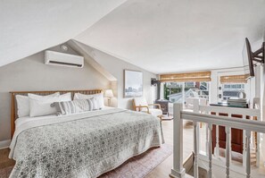 Ein Schlafzimmer mit schrägen Wänden, einem Bett mit gemustertem Überwurf, einem Nachttisch und einem Fenster mit Blick auf ein Haus und Bäume.