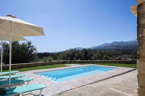 Une piscine rectangulaire, bordée d’une terrasse carrelée, entourée d’un petit muret en pierre et d’une zone herbeuse.