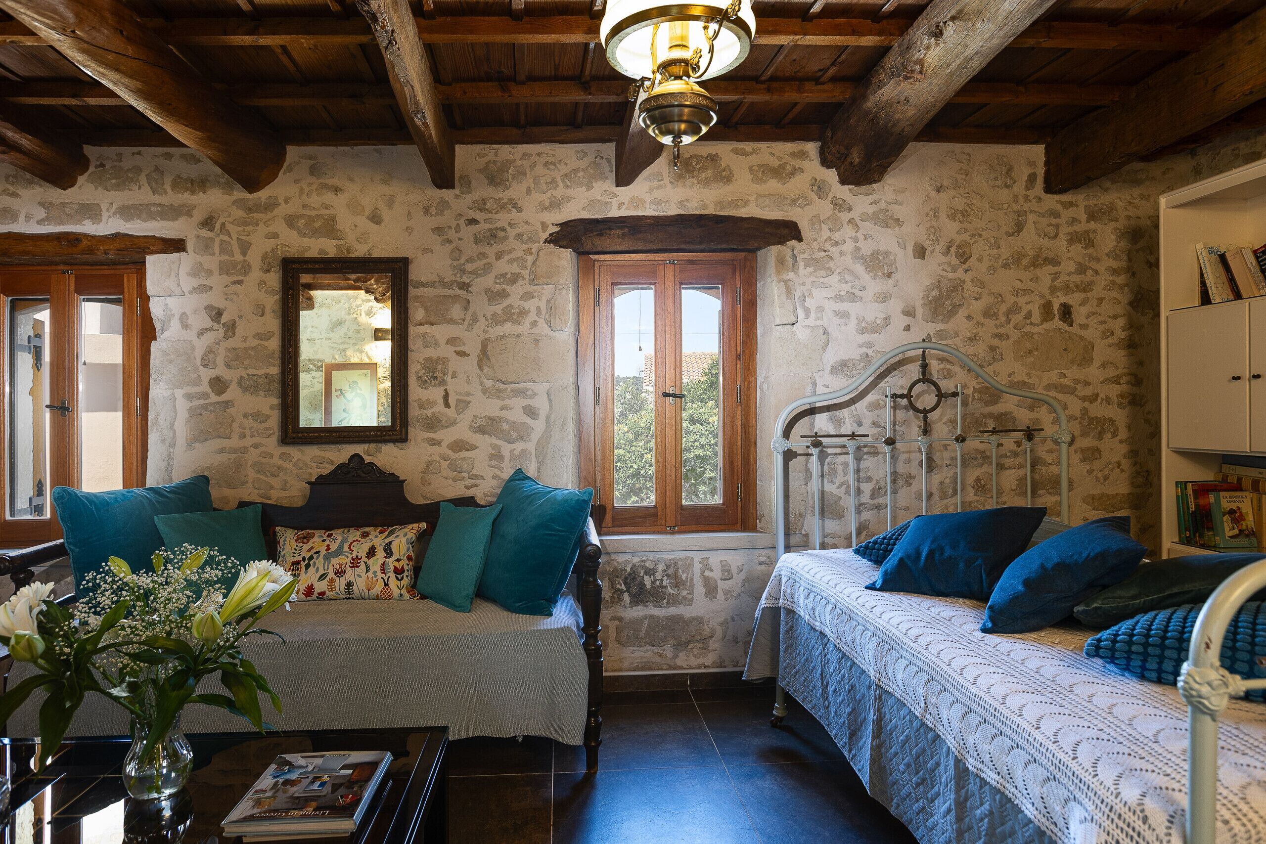 Una stanza rustica con pareti in pietra, travi in legno, un letto con biancheria azzurra, un divano con cuscini color acquamarina e una testiera in legno.