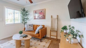Ein Wohnzimmer mit einer Couch, einem Couchtisch, Pflanzen, einem Gemälde und einem Fernseher.