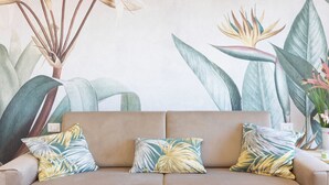 Ein beiges Sofa mit drei tropisch gestalteten Kissen, vor einer Wand mit großen botanischen Drucken.