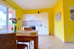 Un interior amarillo brillante con una mesa de comedor de madera y sillas de mimbre, armarios blancos y una zona de cocina con electrodomésticos blancos.