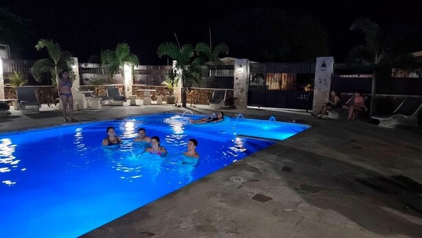 A swimming pool area at night with people in the water, lounge chairs, and a stone wall in the background.
