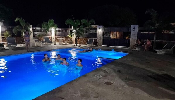 A swimming pool area at night with people in the water, lounge chairs, and a stone wall in the background.