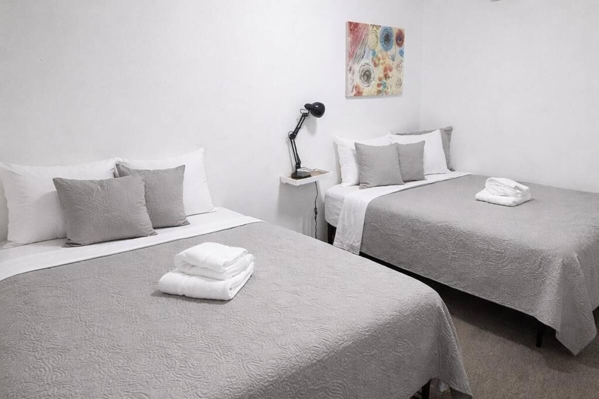 A hotel room with two beds, a nightstand, a lamp, an air conditioner, and a colorful abstract painting on the wall.