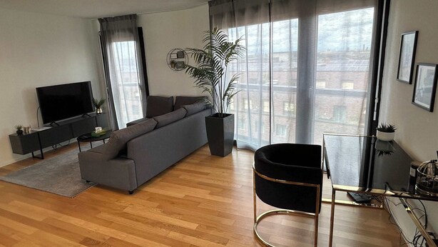 A modern living room with a grey sofa, a black TV stand, a black chair, and a large window with curtains. - Stylish 2.5-Room + Basel Card | 6th-Floor Comfort (Basel)