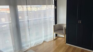 A room with a large window, a chair, and a dark wardrobe. - Stylish 2.5-Room + Basel Card | 6th-Floor Comfort (Basel)