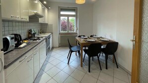 A modern kitchen with white cabinets, a dining table set for four, and a window with a view of trees. - 1st Central 2 BR w/ Living room + 5mn from Basel (Saint-Louis)