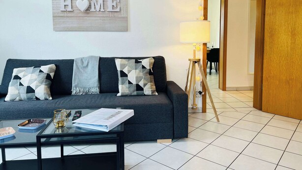 A modern living room with a grey sofa, patterned cushions, a glass coffee table, and a 'HOME' sign on the wall. - 1st Central 2 BR w/ Living room + 5mn from Basel (Saint-Louis)