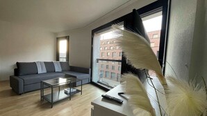 A modern living room with a grey sofa, a glass coffee table, and a view of a brick building through large windows. - Perfect connected to Downtown, Airport & Highway (Basel)