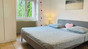 Une chambre à coucher comprenant un lit, un radiateur, une fenêtre, une table de chevet avec une lampe et un tableau accroché au mur.
