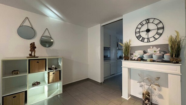A modern living room with a white fireplace, a large wall clock, and a kitchen visible through an open door. - Modern 3 bedroom apartment near ISB and Basel (Reinach)