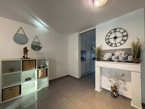 Ein modernes Wohnzimmer mit weißem Kamin, einer großen Wanduhr und einer durchgehenden Tür erkennbaren Küche.