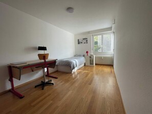 Ein Schlafzimmer mit Holzboden, einem Schreibtisch mit Stuhl, einem Bett, einem Fenster mit Jalousien und einer Heizung.