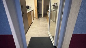 A narrow hallway leading to a kitchen with a sliding glass door, a black mat at the entrance, and a tiled floor.