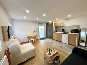 Een knus woongedeelte met een bank, een eettafeltje, een keukenhoek met een magnetron, en een televisie aan de muur.