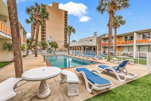 A pool area with lounge chairs and a table, surrounded by palm trees and a building with balconies. - Holiday Shores King Bed (Myrtle Beach)