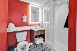 A modern bathroom with a red accent wall, a white toilet, a sink with a mirror above it, and a glass shower enclosure. - Holiday Shores King Bed (Myrtle Beach)