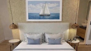 Een slaapkamer met een groot bed, een hoofdeinde met een schilderij van een zeilboot, twee nachtkastjes met lampen, en een houten vloer.