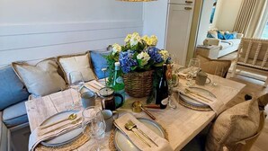 Een eetgedeelte met een lange houten tafel, gedekt voor vier personen, met geweven placemats, gouden bestek en een bloemenstuk als decoratie.