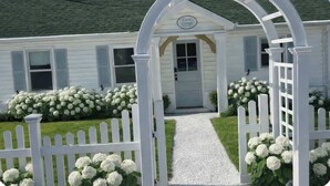 Een wit vakantiehuisje met een veranda, een witte picket fence en witte bloemen.