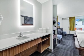 Ein modernes Hotelzimmer mit einem weißen Schminktisch, einem großen Spiegel, einem Holzschrank, einem Bett, einem Schreibtisch und Blick auf die Stadt durch das Fenster.