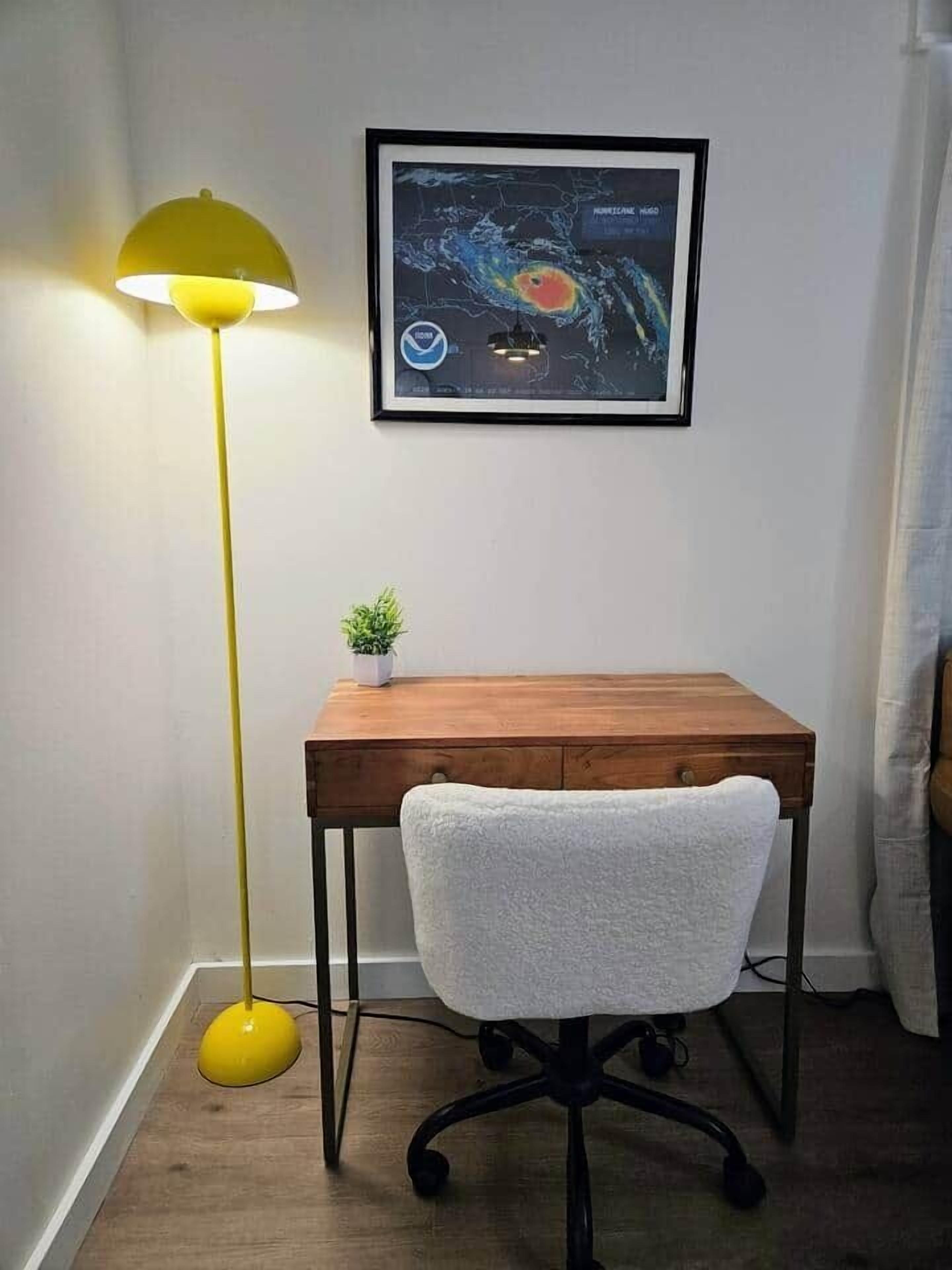 Ein Eckzimmer mit einem Holztisch, einem weißen Stuhl, einer gelben Stehlampe und einer gerahmten Karte eines Hurrikans an der Wand.