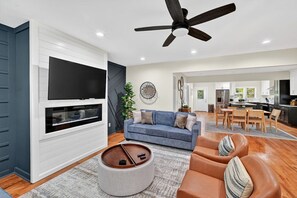 A modern living room with a flat-screen TV, a ceiling fan, a blue sofa, a brown leather armchair, and a dining area in the background. - Grove Park Haven Sleeps 12, Large Backyard (Atlanta)