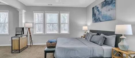 A bedroom with a bed, two bedside tables with lamps, a desk with a lamp, a ceiling fan, and a large window with blinds.