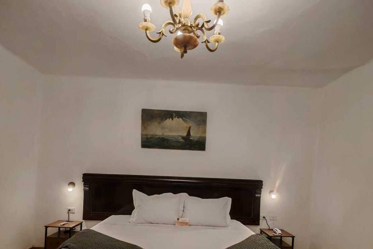 A bedroom with a large bed, wooden flooring, a chandelier, bedside lamps, and a painting on the wall.
