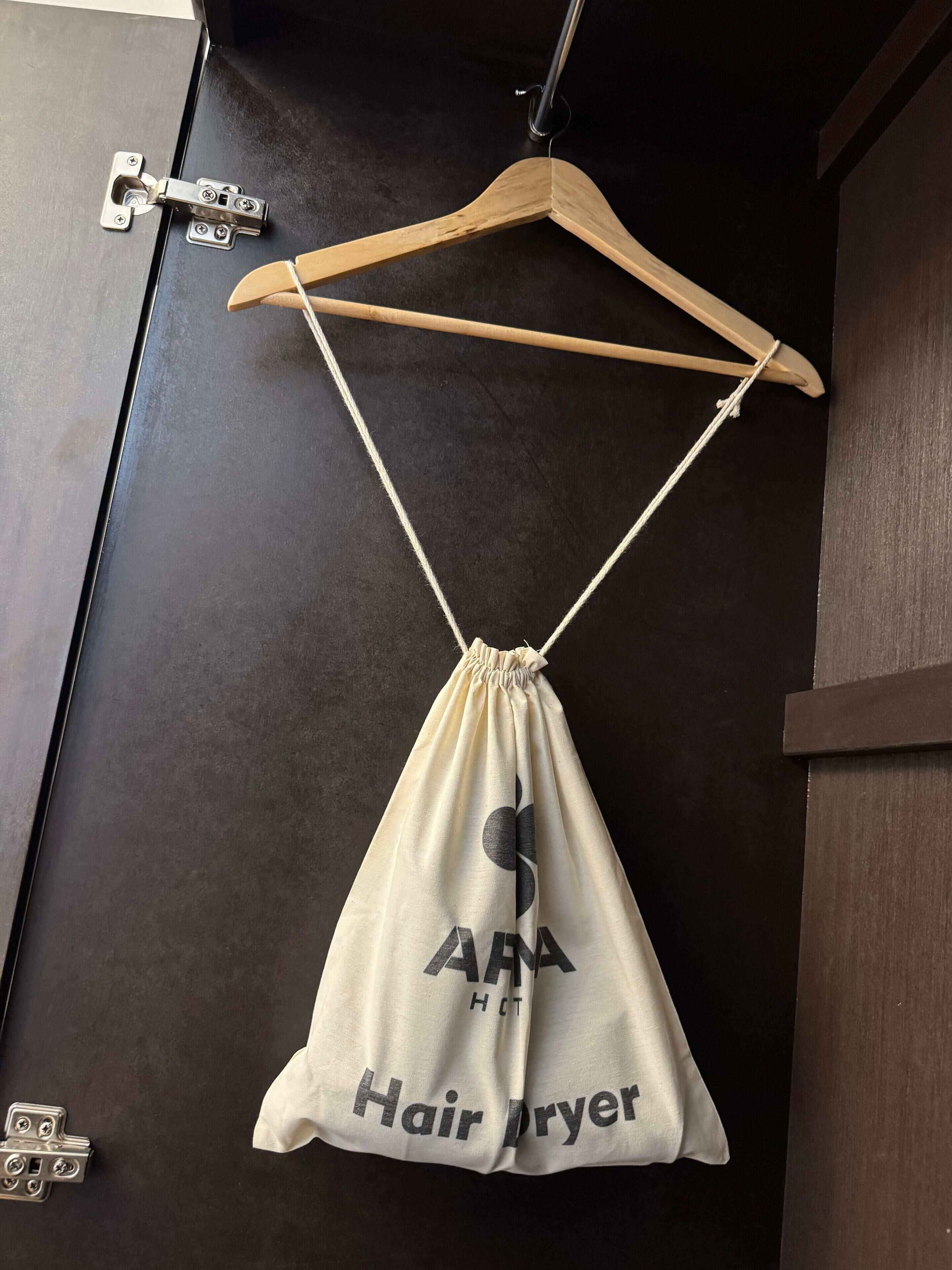 Uma bolsa de tecido pendurada com a inscrição "AAA Hair Dryer", um cabide de madeira e uma tranca de metal numa porta escura.