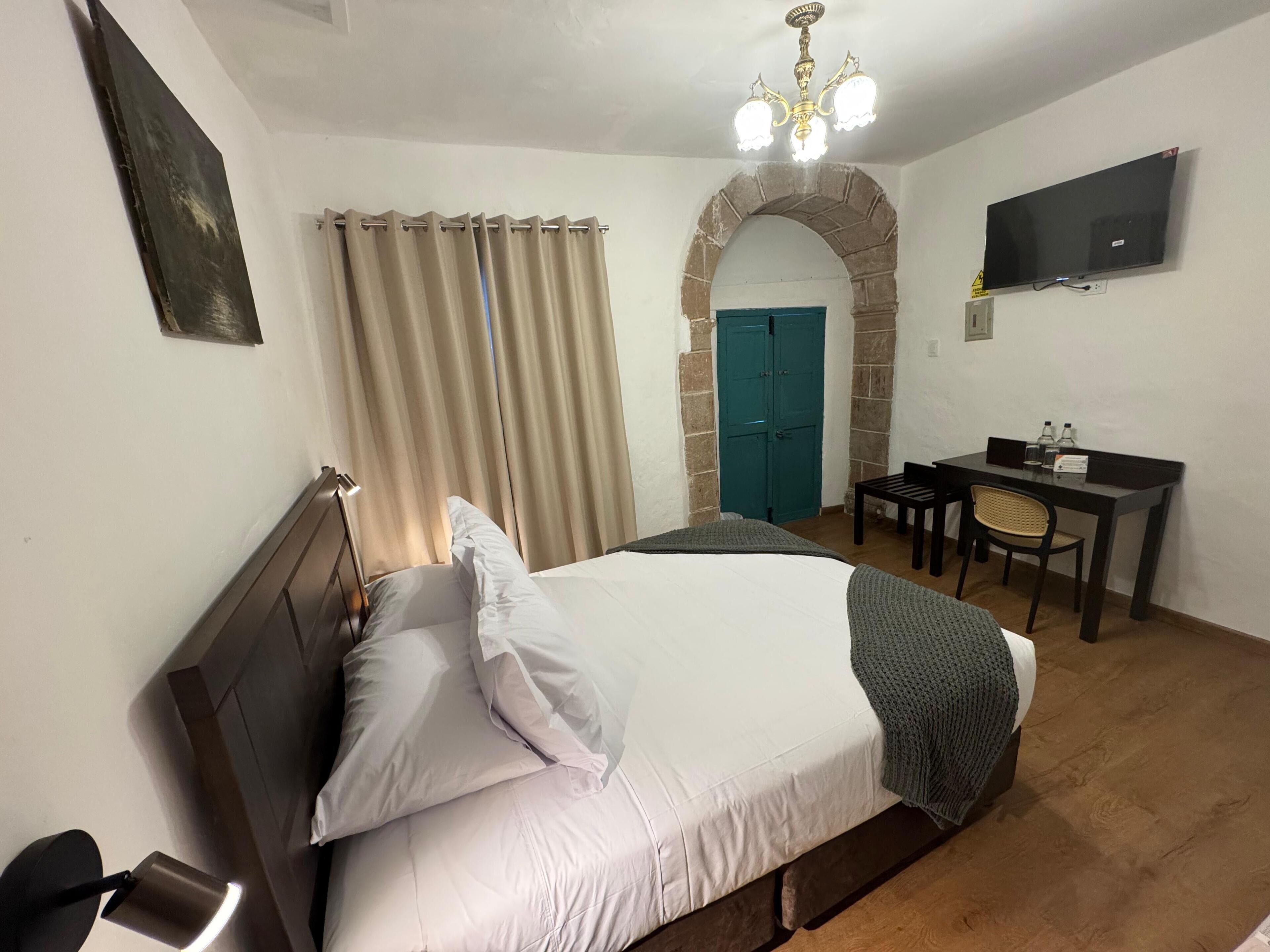 A bedroom with a bed, a desk, a chair, a television, and a stone archway.